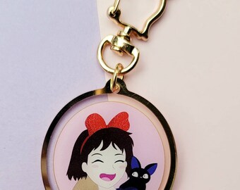 Può includere: Un portachiavi color oro con un ciondolo circolare con un'illustrazione a cartoni animati di una ragazza sorridente con un fiocco rosso e un gatto nero. Il portachiavi ha una chiusura a forma di gatto.