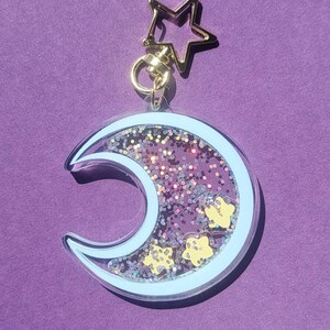Pu&ograve; includere: Un portachiavi a forma di mezzaluna con un contorno blu chiaro e glitter all'interno. Il glitter &egrave; rosa, viola e oro. Ci sono piccole stelle gialle all'interno del glitter. Il portachiavi ha un anello dorato a forma di stella nella parte superiore.