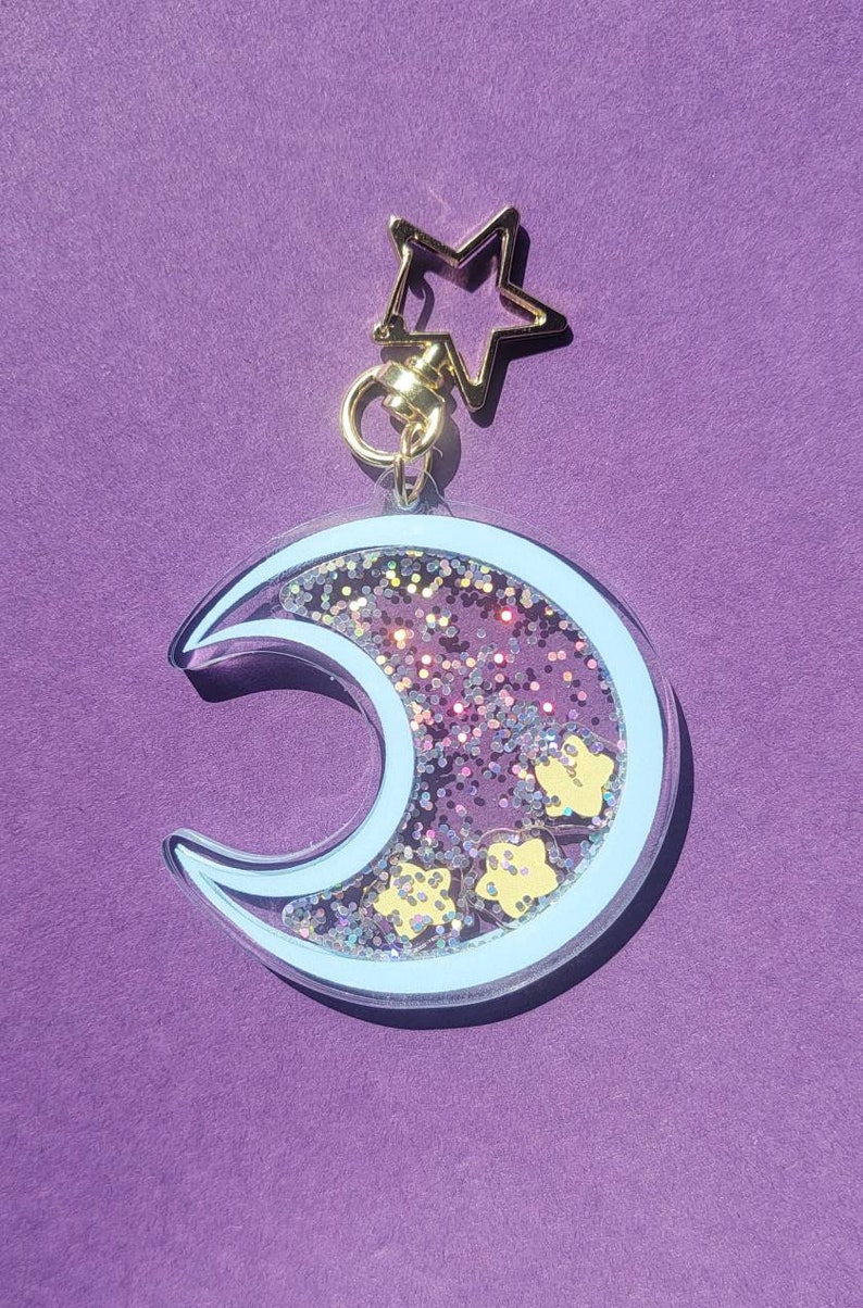 Pu&ograve; includere: Un portachiavi a forma di mezzaluna con un contorno blu chiaro e glitter all'interno. Il glitter &egrave; rosa, viola e oro. Ci sono piccole stelle gialle all'interno del glitter. Il portachiavi ha un anello dorato a forma di stella nella parte superiore.