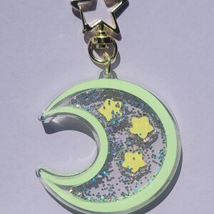 Pu&ograve; includere: Un portachiavi a forma di mezzaluna verde con glitter e stelle gialle all'interno. Il portachiavi ha un fermaglio a forma di stella dorata.