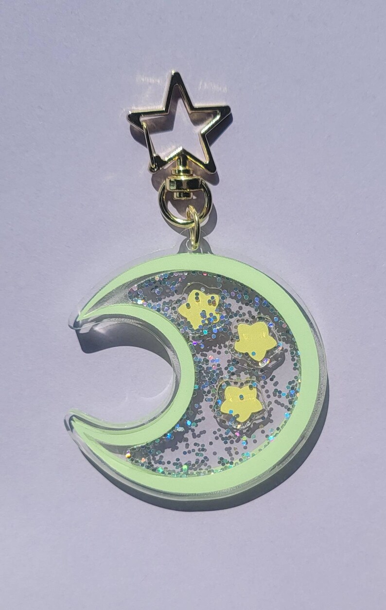 Pu&ograve; includere: Un portachiavi a forma di mezzaluna verde con glitter e stelle gialle all'interno. Il portachiavi ha un fermaglio a forma di stella dorata.