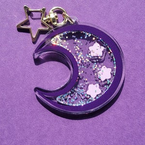 Pu&ograve; includere: Un portachiavi a forma di luna crescente viola con un ciondolo a stella dorata. La luna &egrave; piena di glitter iridescente e stelle bianche.