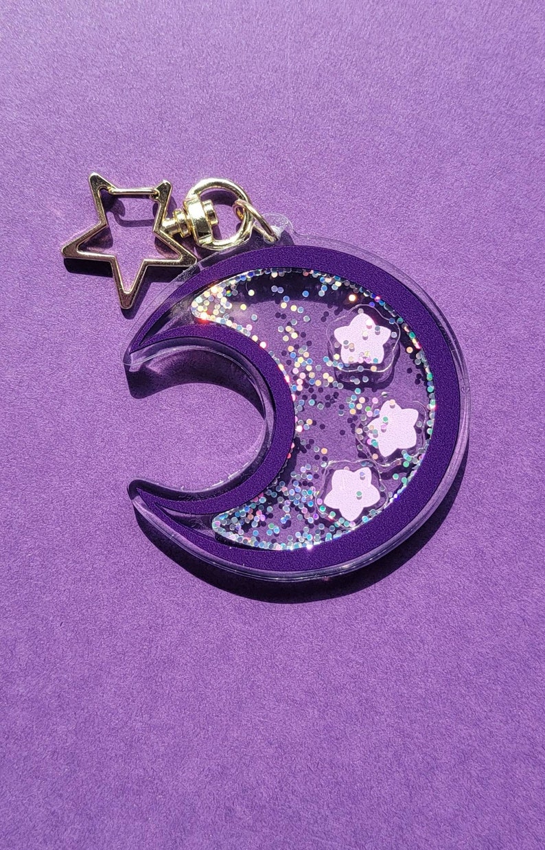 Pu&ograve; includere: Un portachiavi a forma di luna crescente viola con un ciondolo a stella dorata. La luna &egrave; piena di glitter iridescente e stelle bianche.
