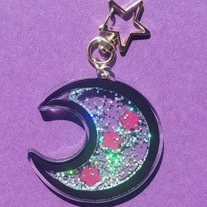 Pu&ograve; includere: Un portachiavi a forma di mezzaluna con un contorno nero e un interno in resina trasparente riempito di glitter argentati e coriandoli a forma di fiori rosa. Un ciondolo a forma di stella dorata &egrave; attaccato alla parte superiore del portachiavi.