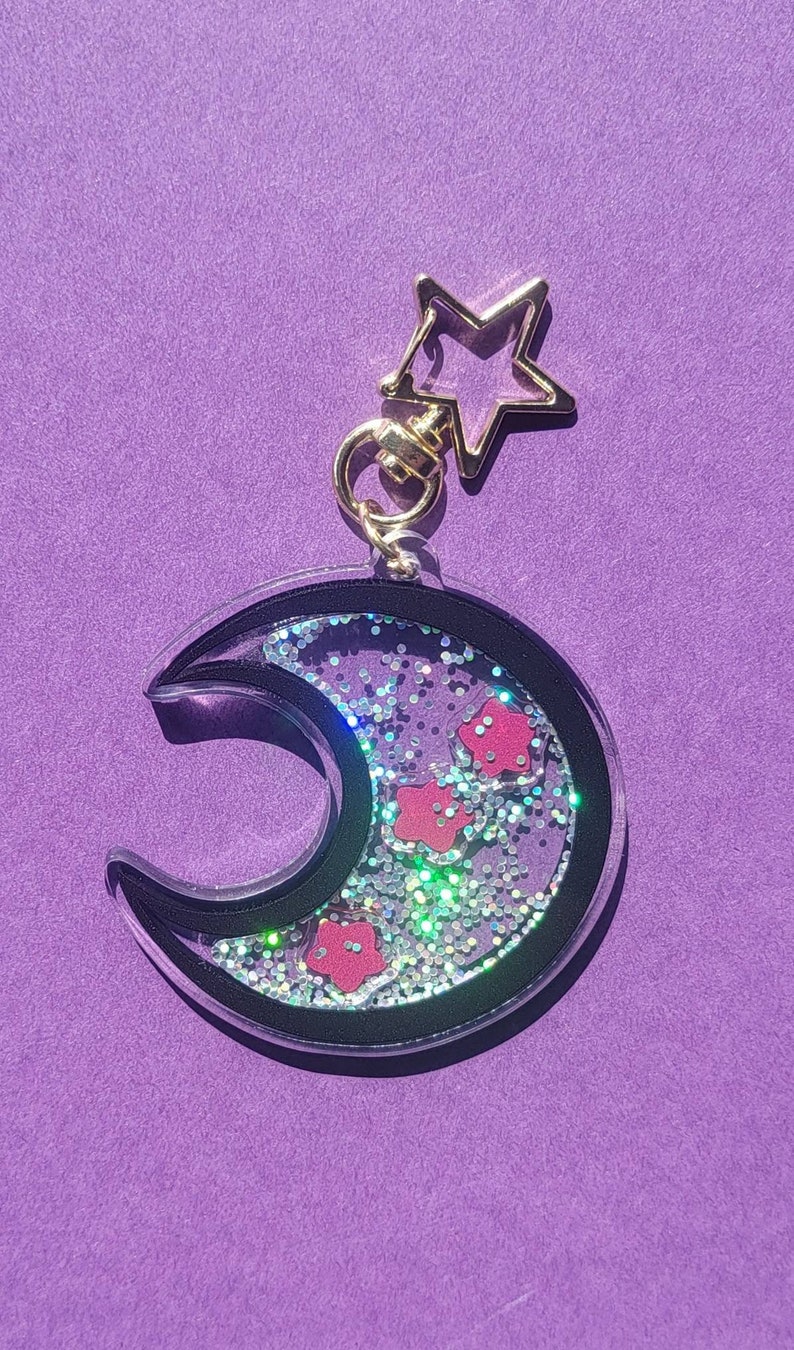 Pu&ograve; includere: Un portachiavi a forma di mezzaluna con un contorno nero e un interno in resina trasparente riempito di glitter argentati e coriandoli a forma di fiori rosa. Un ciondolo a forma di stella dorata &egrave; attaccato alla parte superiore del portachiavi.