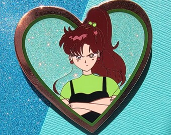 Può includere: Uno spillo a forma di cuore con un bordo verde e marrone. Lo spillo presenta un personaggio dei cartoni animati con lunghi capelli rossi, che indossa una camicia verde e un vestito nero, con un'espressione seria.