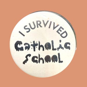Könnte beinhalten: Ein runder, cremefarbener Button mit dem Text "I SURVIVED Catholic School" in schwarzer, stilisierter Schrift. Der Button hat einen silbernen Rand und einen pfirsichfarbenen Hintergrund.