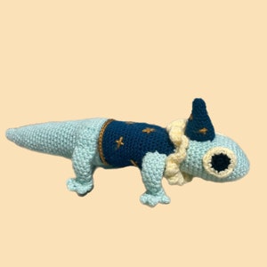 Peut inclure: Un lézard crocheté bleu et blanc portant un gilet à motifs d'étoiles bleues et dorées et un chapeau jaune et blanc.