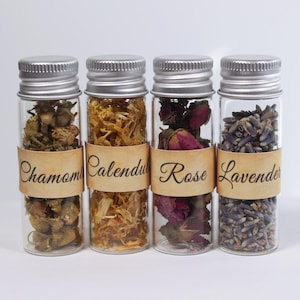 May include: Four small glass bottles with silver lids, each filled with dried herbs and flowers. The bottles are labeled with the names: Chamomile, Calendula, Rose, and Lavender. The labels are a light brown color.