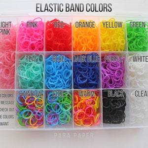 May include: A clear plastic storage container filled with small elastic bands in various colors. The compartments are labeled with text including Light Pink, Pink, Red, Orange, Yellow, Green, Teal, Blue, Dark Blue, Purple, White, Rainbow, Cool, Warm, Black, and Clear.