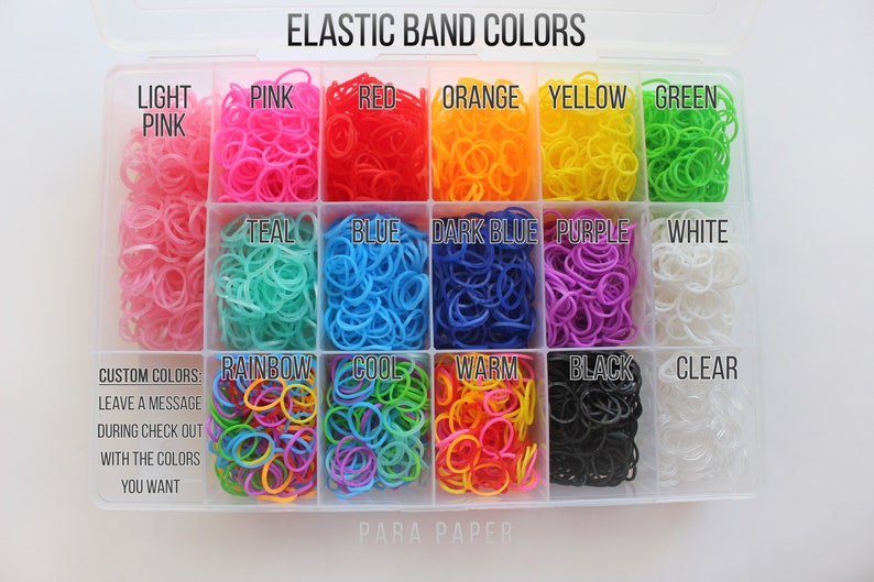 May include: A clear plastic storage container filled with small elastic bands in various colors. The compartments are labeled with text including Light Pink, Pink, Red, Orange, Yellow, Green, Teal, Blue, Dark Blue, Purple, White, Rainbow, Cool, Warm, Black, and Clear.