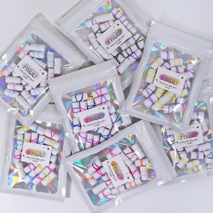 May include: Clear plastic bags filled with small, white paper rolls wrapped with colorful rubber bands. Each bag has a small card with the word "PRIDE!" and text. The bags are arranged on a white surface, with a holographic sheen.