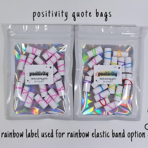 May include: Two clear holographic pouches containing small white paper rolls secured with elastic bands. The left pouch features a white label with the text 'positivity twenty inspiring quotes para paper' and pink bands. The right has a rainbow label and multicolored bands.