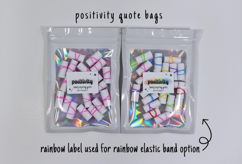 May include: Two clear holographic pouches containing small white paper rolls secured with elastic bands. The left pouch features a white label with the text 'positivity twenty inspiring quotes para paper' and pink bands. The right has a rainbow label and multicolored bands.