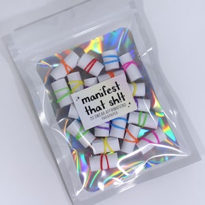 May include: A clear plastic bag filled with small white paper scrolls tied with colorful rubber bands. The bag has a holographic backing and a white label with the text "manifest that sh!t" and "20 swear affirmations".