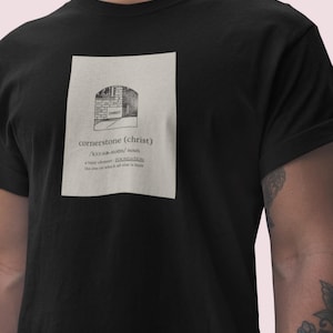 Pode incluir: Camiseta preta com um gráfico branco de um edifício de tijolo com a palavra "CHRIST" nele. O gráfico também inclui o texto "cornerstone (christ)" e uma definição da palavra cornerstone.