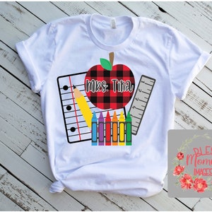 K&ouml;nnte beinhalten: Wei&szlig;es T-Shirt mit einem Design zum Thema Schule. Das Design umfasst einen rot-schwarz karierten Apfel, einen Notizblock, einen gelben Bleistift, ein Lineal und einen Satz farbiger Buntstifte. Der Text "Mrs. Tika" steht auf dem Apfel.