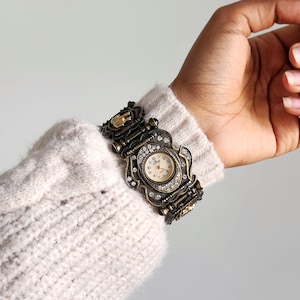 May include: An antique-style watch with a bronze-colored metal band and face. The watch face is cream-colored with black numerals and hands. The watch is adorned with small, clear crystals. The watch is worn on a wrist covered by a cream-colored sweater.