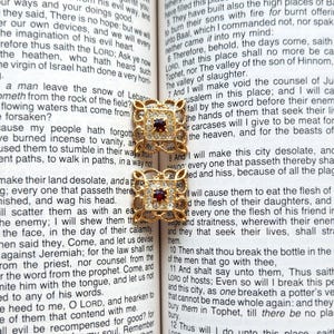 May include: Two gold-toned earrings with a square design, featuring a central red gemstone and surrounded by smaller clear stones. The earrings are set against the backdrop of an open book.