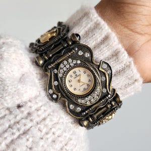 May include: Ornate antique-style watch with a bronze-tone metal band and case. The watch face is cream-colored with black numerals and hands, surrounded by sparkling crystals. The watch is a unique accessory.
