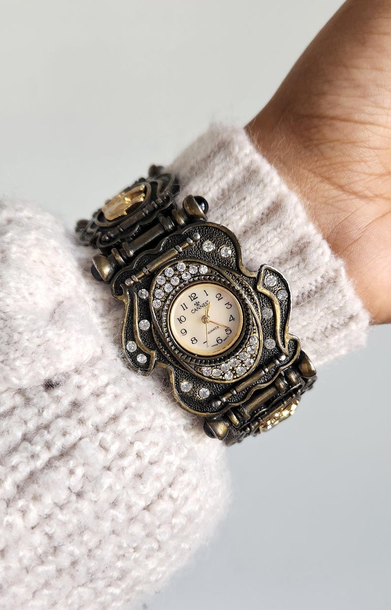 May include: Ornate antique-style watch with a bronze-tone metal band and case. The watch face is cream-colored with black numerals and hands, surrounded by sparkling crystals. The watch is a unique accessory.