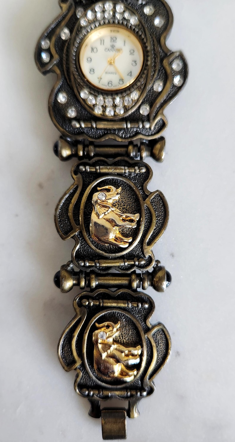May include: Vintage gold-toned watch with a decorative band. The watch face is surrounded by clear crystals. The band features gold elephant figures set in ornate frames. The watch has a quartz movement.