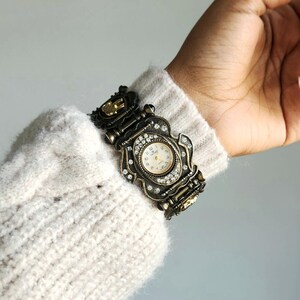 May include: Ornate antique-style wristwatch with a bronze-colored metal band and case. The watch face is cream-colored with black numerals and hands. The case is embellished with clear crystals. The watch is worn on a wrist covered by a cream-colored sweater.