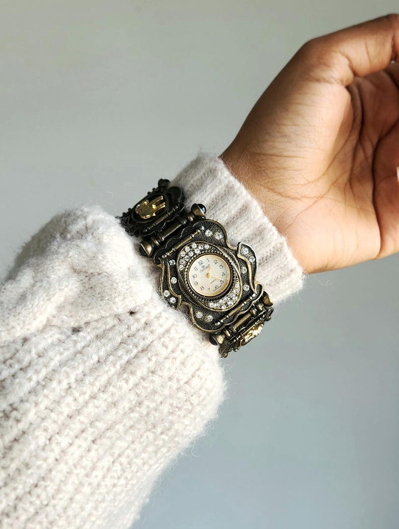 May include: Ornate antique-style wristwatch with a bronze-colored metal band and case. The watch face is cream-colored with black numerals and hands. The case is embellished with clear crystals. The watch is worn on a wrist covered by a cream-colored sweater.