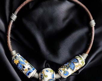 May include: A brown leather necklace with silver wire accents and three ceramic beads. The beads are white with blue and yellow floral patterns.