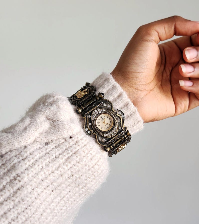 May include: An antique-style watch with a bronze-colored metal band and face. The watch face is cream-colored with black numerals and hands. The watch is adorned with small, clear crystals. The watch is worn on a wrist covered by a cream-colored sweater.