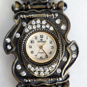 May include: Vintage gold-toned watch with a decorative band. The watch face is surrounded by clear crystals and features the word "Cannes". The band has ornate details and gold-colored accents.