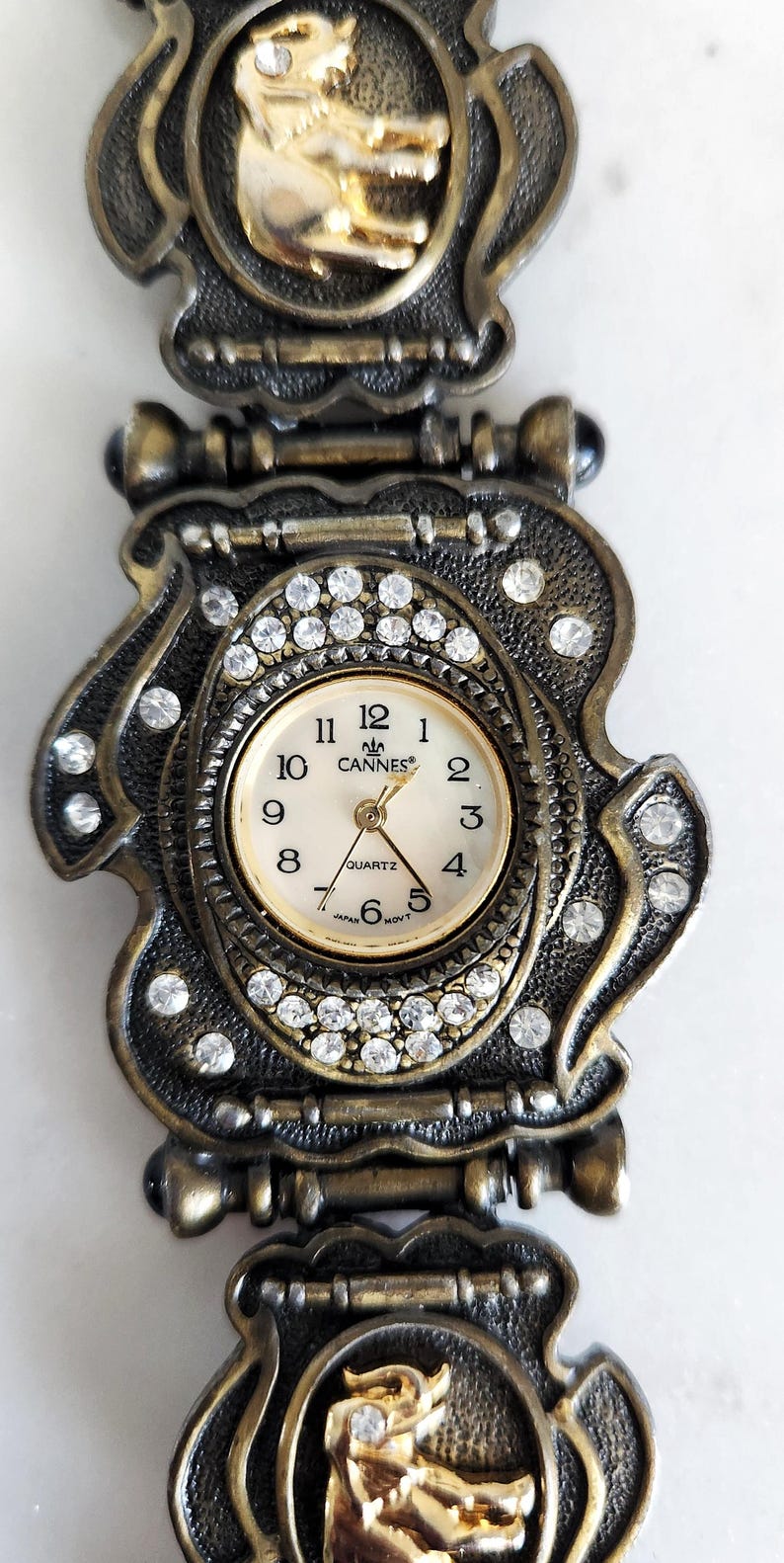 May include: Vintage gold-toned watch with a decorative band. The watch face is surrounded by clear crystals and features the word "Cannes". The band has ornate details and gold-colored accents.