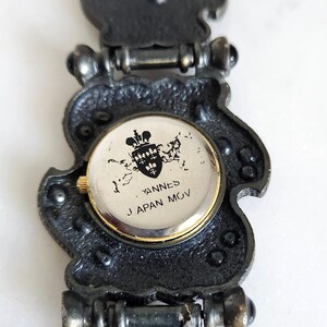 May include: Close-up of a vintage watch with a decorative, dark metal band. The watch face is round with gold trim and features the text "YANNES JAPAN MOV" and a crest. The band has an ornate, textured design.
