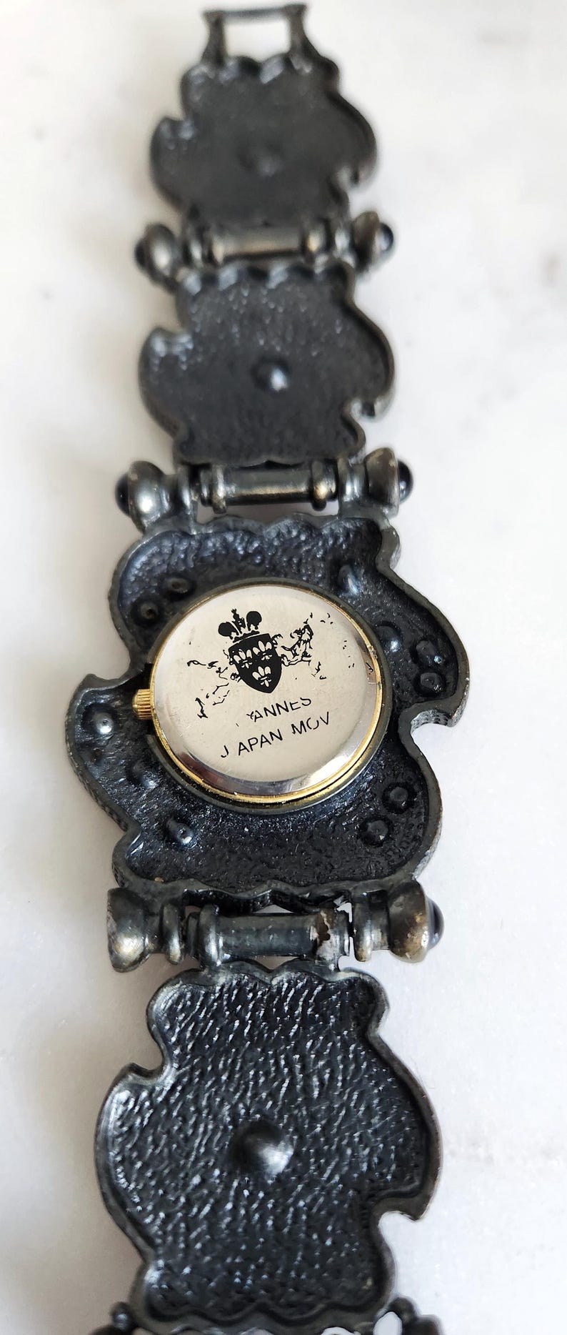 May include: Close-up of a vintage watch with a decorative, dark metal band. The watch face is round with gold trim and features the text "YANNES JAPAN MOV" and a crest. The band has an ornate, textured design.