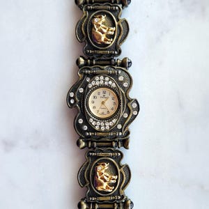 May include: Vintage gold-toned watch with a decorative band featuring ornate frames and gold-colored bear figures. The watch face is surrounded by rhinestones and has a cream-colored dial with black numerals.