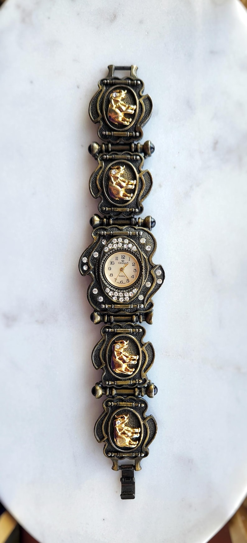 May include: Vintage gold-toned watch with a decorative band featuring ornate frames and gold-colored bear figures. The watch face is surrounded by rhinestones and has a cream-colored dial with black numerals.