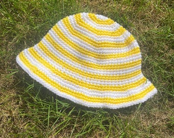 May include: A crochet bucket hat featuring horizontal stripes in bright yellow and white. The hat has a rounded crown and a downward-sloping brim, constructed with a consistent stitch pattern throughout the entire piece.