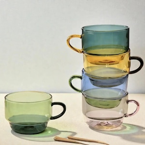 Può includere: Una pila di quattro tazze in vetro traslucido nei colori verde acqua, ambra, blu e verde chiaro, con una singola tazza in vetro verde chiaro posizionata a sinistra. Ogni tazza presenta un manico semplice e una forma cilindrica liscia.