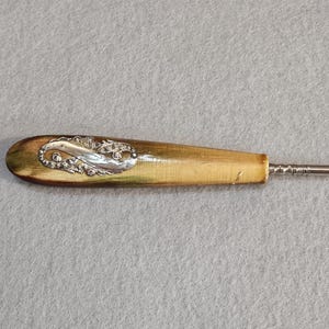 May include: A vintage crochet hook with a wooden handle and a decorative silver-toned embellishment. The hook is made of metal and has a curved end. The handle is light brown with a silver design.