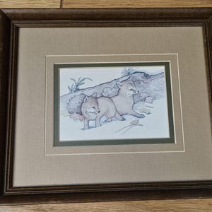 May include: A framed print featuring a line drawing of two fox kits resting near a fallen log. The artwork is set within a double matting of beige and dark green, enclosed in a dark brown wooden frame with a textured finish and visible grain.