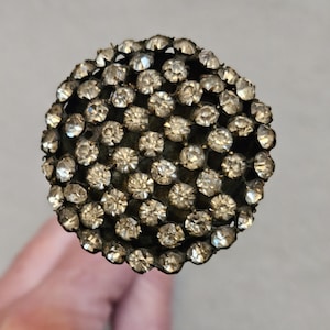 May include: A round, dark-toned object densely covered with small, sparkling, clear crystals. The crystals are evenly spaced, creating a textured, glittering surface. The object appears to be a decorative element, possibly a knob or finial.
