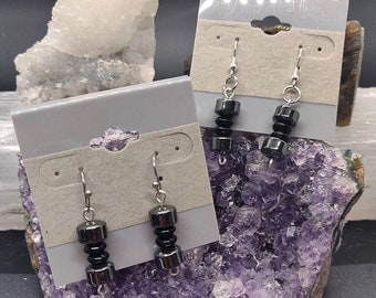 May include: A pair of black dumbbell-shaped earrings with silver hooks. The earrings are displayed on a white card with a hole for hanging.