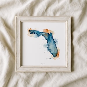 Puede incluir: Una impresión en acuarela enmarcada del estado de Florida. La obra de arte presenta un diseño de acuarela azul y naranja con la forma del estado, con la palabra "Florida" impresa en la parte inferior. El marco es de color claro y neutro.