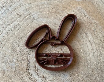 May include: Brown plastic cookie cutter shaped like a bunny wearing a face mask. The bunny has large ears, a round face, and a small nose. The face mask has a striped pattern.
