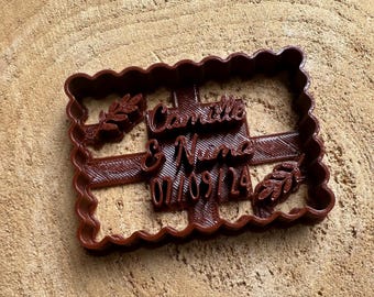 May include: A brown plastic cookie cutter shaped like a rectangle with a white cross pattern. The cutter has the text "Camille & Nima 07.09.14" and two small leaf designs.