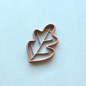 Peut inclure: Un emporte-pièce en métal couleur cuivre en forme de feuille de chêne. Le découpoir présente un contour détaillé de la forme de la feuille, y compris les nervures. Le fond est de couleur bleu clair.