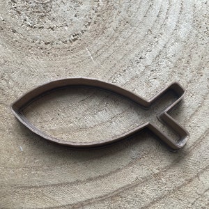 May include: A brown plastic cookie cutter shaped like a fish. The cutter is on a light brown wooden surface.