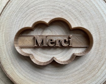 May include: A brown plastic cookie cutter shaped like a cloud with the word "Merci" in raised letters.
