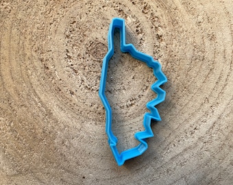 May include: A light blue plastic cookie cutter in the shape of a geographical outline, possibly a state or region. The cutter is placed on a textured, light brown wooden surface.
