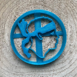 Op de afbeelding: Een blauwe plastic koekjesvormer in de vorm van een mensenhoofd met een hoofdband. De koekjesvormer is rond en heeft een kruisvormig ontwerp in het midden.